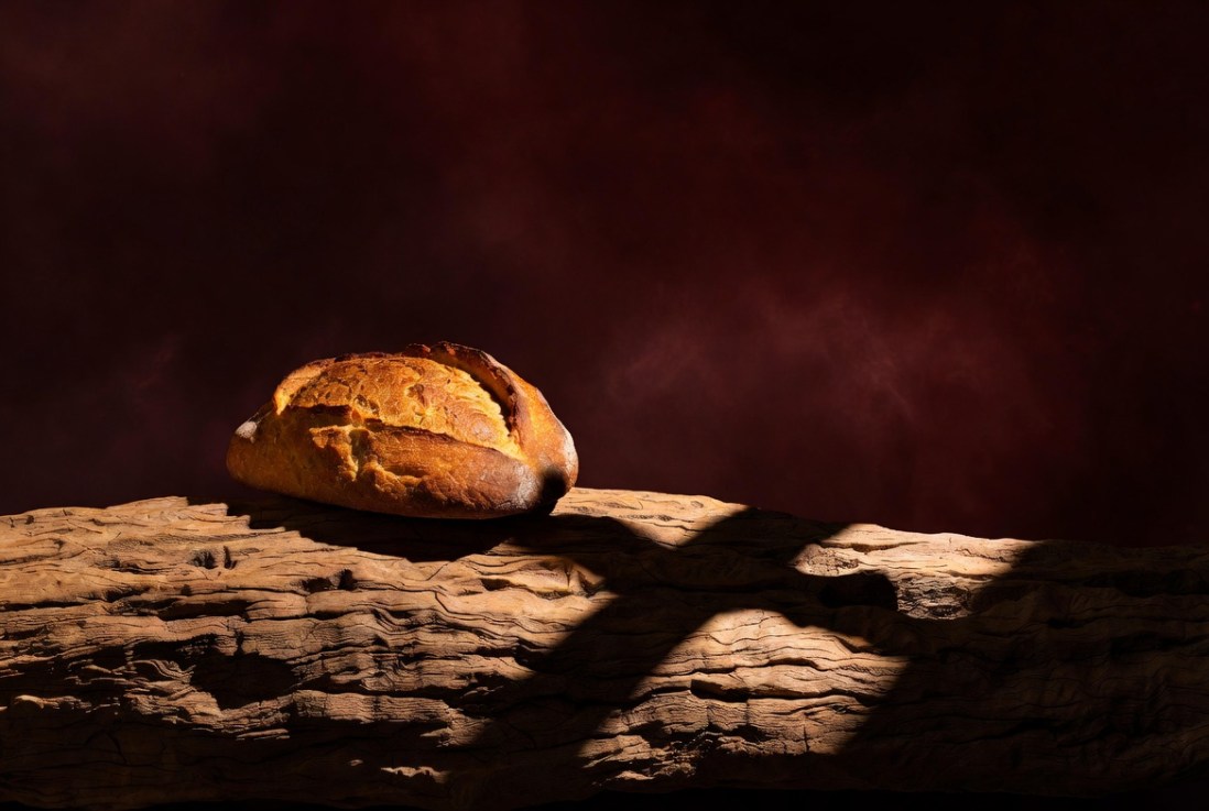 "A loaf of bread on weathered wood, with the shadow of a cross falling across the surface — a visual meditation on Christ as the Bread of Life and the cross as the tree of redemption."